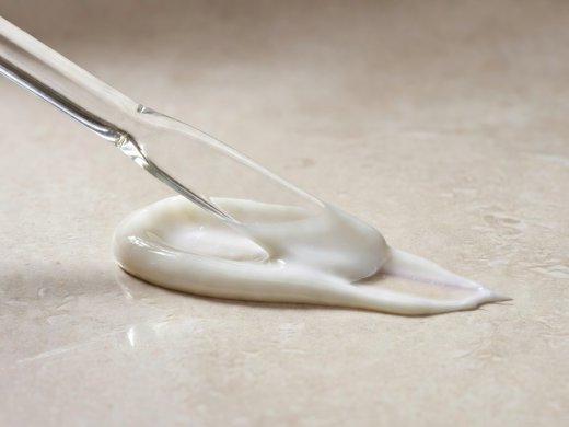 Close-up of a clear glass spatula spreading white emulsion on a warm neutral tile